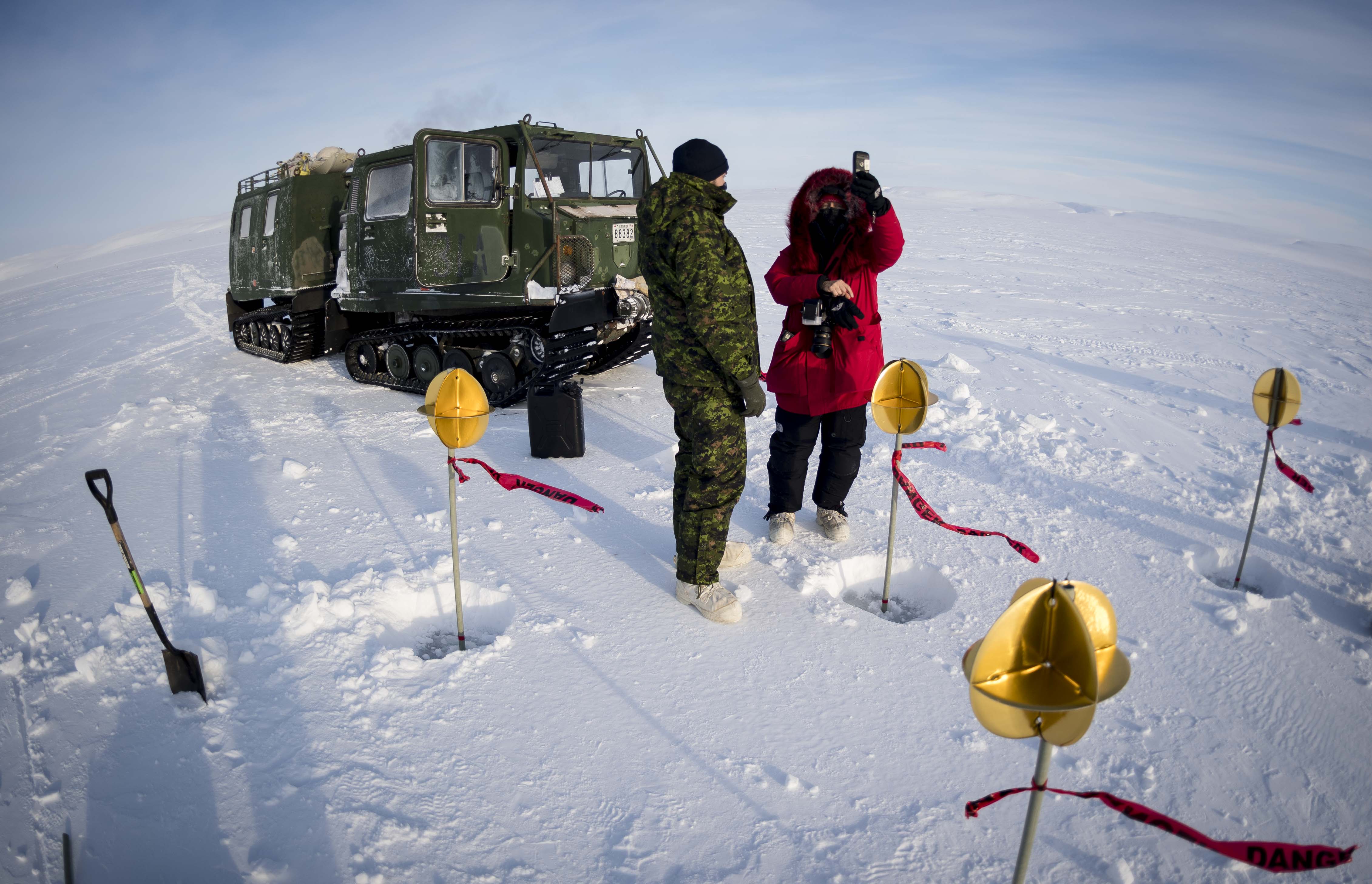 Two people, one wearing a camouflage uniform and the other winter clothing, stand in snow in front of a green tracked vehicle. Around them are small poles planted in the snow with round yellow objects at their tip.