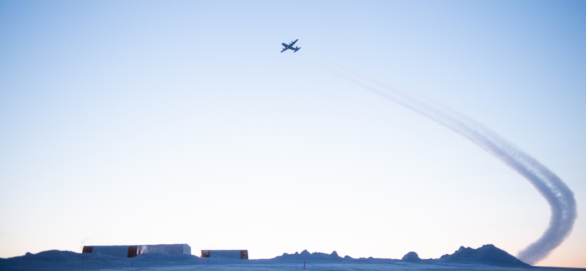 In a photo that shows an Arctic landscape, a big propeller aircraft flies in a blue sky leaving a vapour trail behind it.